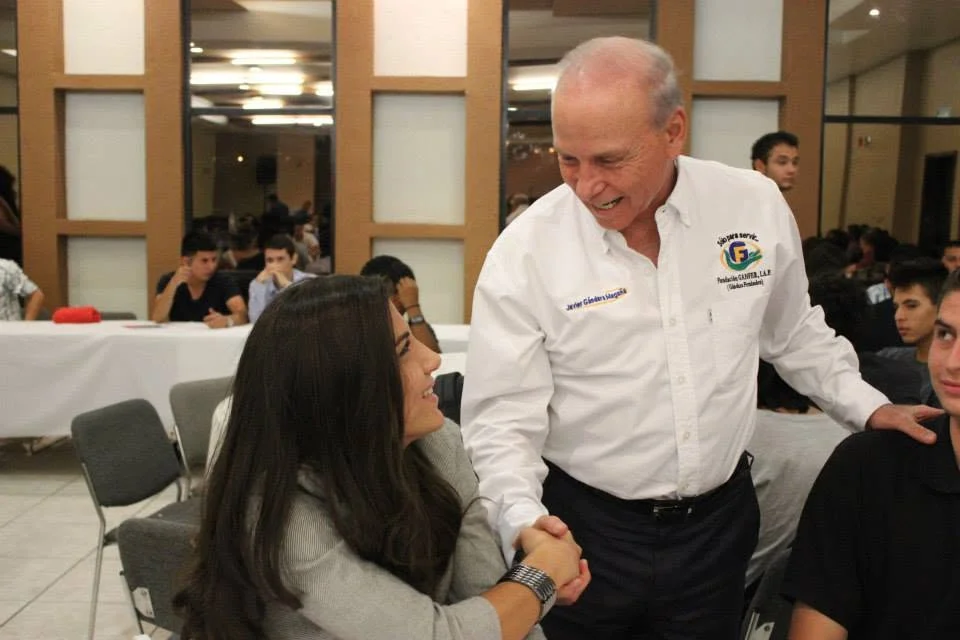Javier Gándara Magaña saluda a una asistente durante un recorrido por un evento de participación comunitaria.